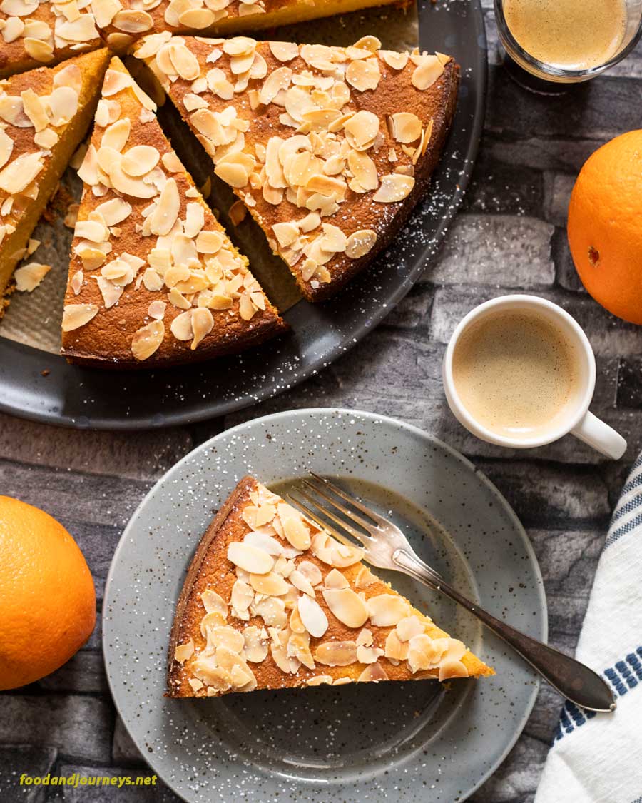 A slice of French Orange and Almond Flourless cake on a plate served with a shot of espresso. Oranges and the rest of the cake on a baking tray are also on the same table.