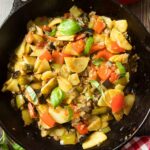An overhead shot of a skillet of potatoes and peppers, with fresh red bell pepper next to it.